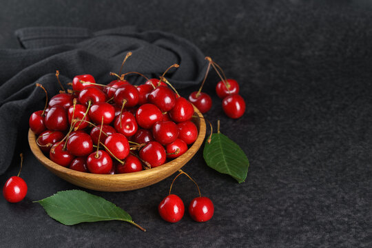 Fresh ripe red sweet cherries in wooden bowl on dark textured stone background with green leaves and black linen towel. Summer fruit, berry, harvest, healthy organic food