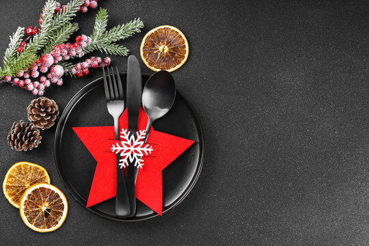 Holiday table setting with black plate and black cutlery in red star shaped cutlery holder with white snowflake and frosted Christmas tree branch on dark textured background, copy space.