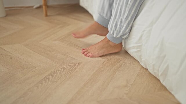 Woman sitting on bed edge tiptoeing with bare feet and painted toenails, pajama trousers, toes touching wooden floor inside a residential building; serenity morning routine.