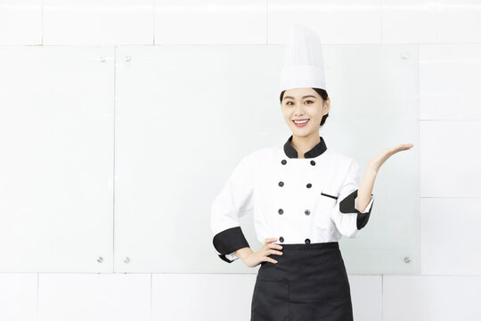 Smiling Female Chef in Uniform Presenting with Hand in Bright Kitchen