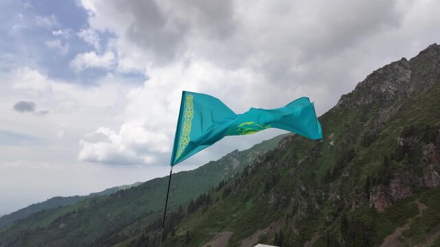 National flag of Kazakhstan waving in breeze against scenic mountain peaks and cloudy sky in Shymbulak, Almaty