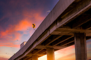 Obraz premium Elevated highway bridge against dramatic orange sunset sky with cargo truck, urban transportation infrastructure, road engineering and freight logistics concept.