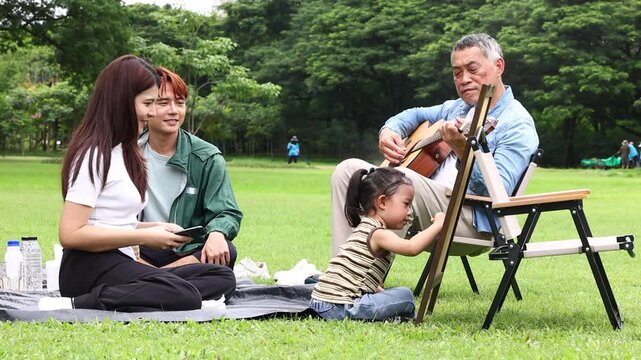 Asian kids painting in park. Asian family enjoy outdoor picnic with food and drinks in park sunny day, outdoor activitie with daughter father, mother grandfather. GrandFather playing acoustic guitar