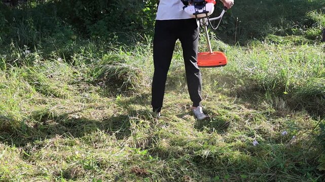 Gardener trimming weeds and overgrown grass with a motor scythe during summer dacha work