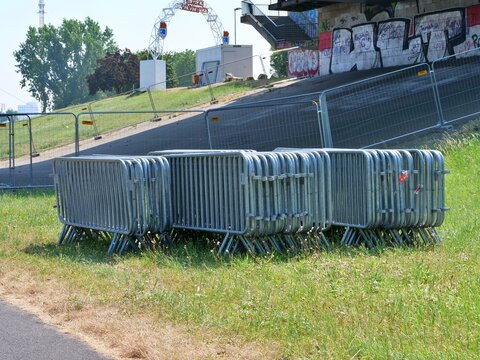 Stacked metal crowd control barriers sitting on a green grass field near a bridge overpass preparing for a large public outdoor festival event.