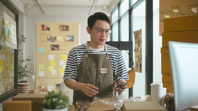 Cheerful Asian male small business owner using smartphone to take a photo of a cardboard box parcel, preparing online customer order for delivery in a home office workspace.
