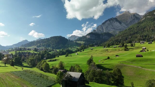 Aerial drone view of green alpine meadows with wooden barns and a village below rocky peaks in Switzerland