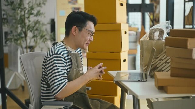 Joyful Asian male entrepreneur smiling and talking to a client during an online video conference on his laptop, sitting among shipping boxes.