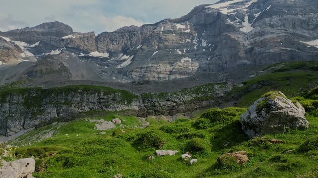 Slow cinematic drone flight just above a lush alpine meadow, moving forward toward an impressive rocky mountain massif. Steep cliffs, patches of snow and vibrant green vegetation create a dramatic hig