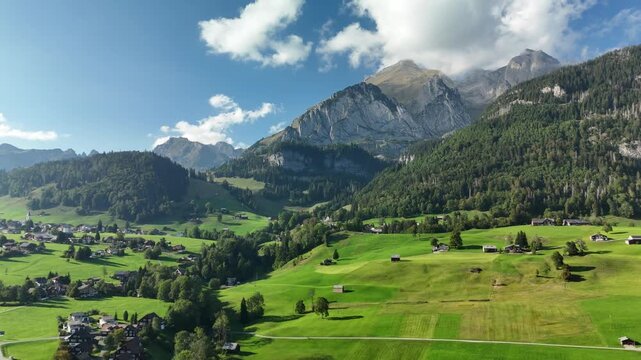 Aerial drone view of green alpine meadows and wooden barns below a rocky mountain ridge in Switzerland