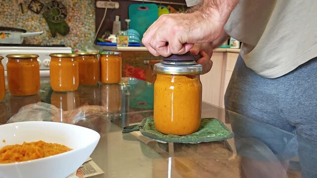 Hands tightening a metal lid on a jar of squash caviar with a canning key on a kitchen table, homemade preserves, traditional recipe.