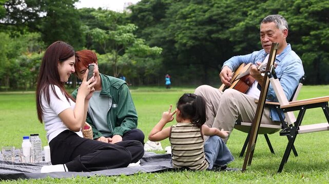 Asian kids painting in park. Asian family enjoy outdoor picnic with food and drinks in park sunny day, outdoor activitie with daughter father, mother grandfather. GrandFather playing acoustic guitar