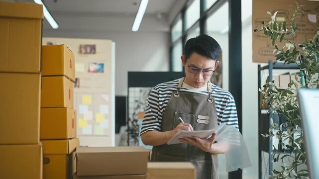 Asian male entrepreneur holding a clipboard and writing notes to check product inventory and manage shipping boxes in his small shop.