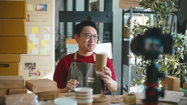 Smiling Asian male entrepreneur presenting sustainable paper cup and wooden spoon recycled cardboard box to the camera, promoting eco-friendly packaging for his small business.