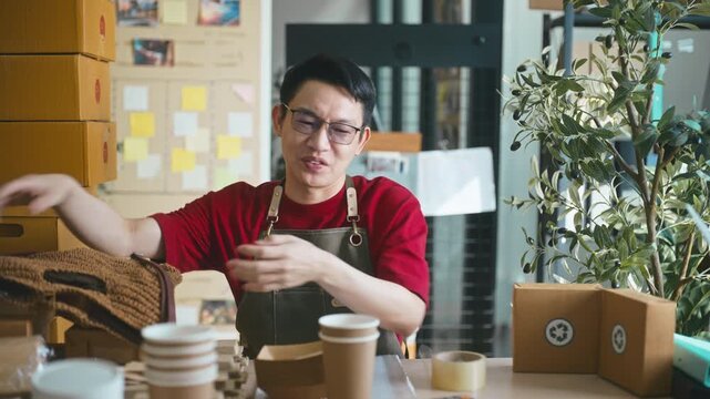Smiling Asian male entrepreneur presenting sustainable paper cup and wooden spoon recycled cardboard box to the camera, promoting eco-friendly packaging for his small business.