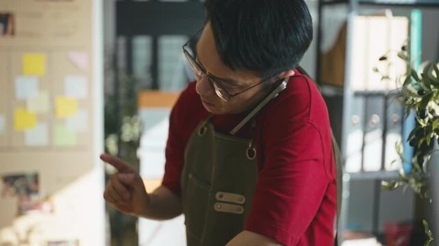 Asian male entrepreneur in a red shirt talking on a smartphone with a customer to check order details.