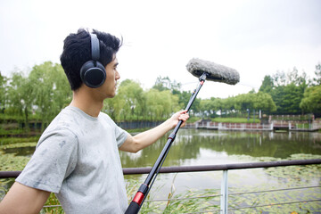 Man Recording Audio with Boom Microphone and Headphones in a Park by a Lake © HaXuanDieu