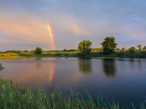 A beautiful double rainbow stretches across the sky, reflecting perfectly on the calm water surface of a scenic rural lake.