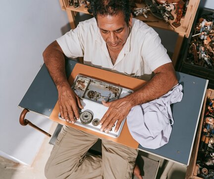 Antique wooden toys and vintage figurines surround a man repairing a weathered wind-up mechanism on a tan tray for collector media and hobbyists