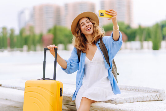 Young female tourist wearing hat records vlog on phone while sitting on bench with yellow suitcase. Woman is relaxing and taking selfie outdoors. Concepts: tourism, blogging, and urban life.