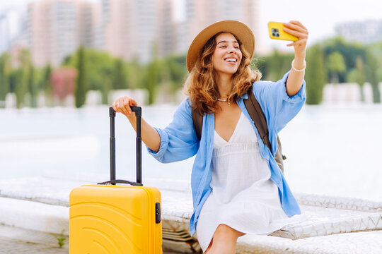 Young female tourist wearing hat records vlog on phone while sitting on bench with yellow suitcase. Woman is relaxing and taking selfie outdoors. Concepts: tourism, blogging, and urban life.