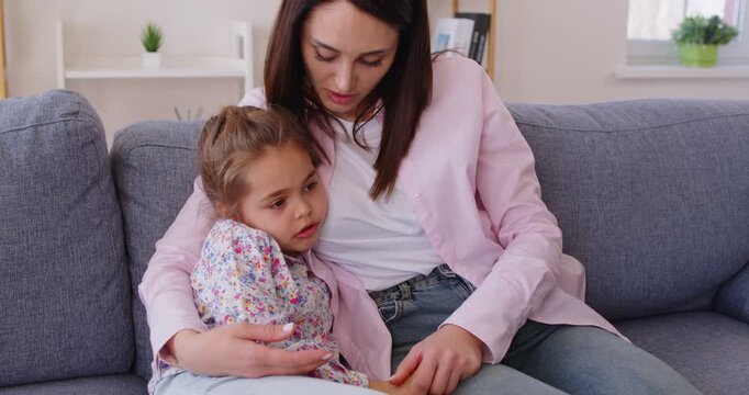 Loving mother cuddling and holding hands with her young daughter on sofa in living room. Caring smiling woman embracing little girl and listening as sad child sharing thoughts, feelings and concerns.