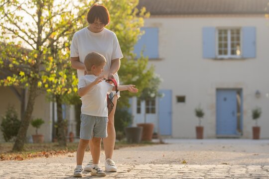 mother and child standing together on cobblestone path in calm countryside house setting with blue shutters and potted plants, side view, white shirts, domestic mood.