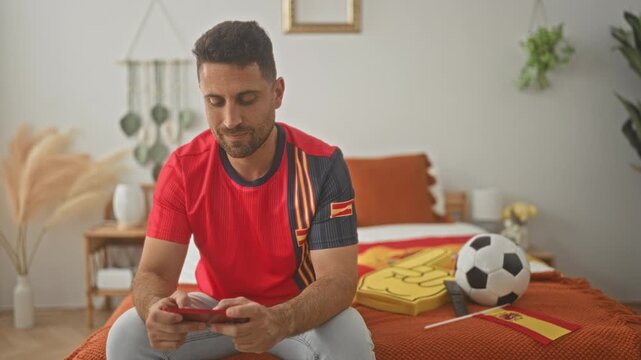 Man on bed holding smartphone and fist pumping while a soccer ball lies nearby inside building; match excitement.