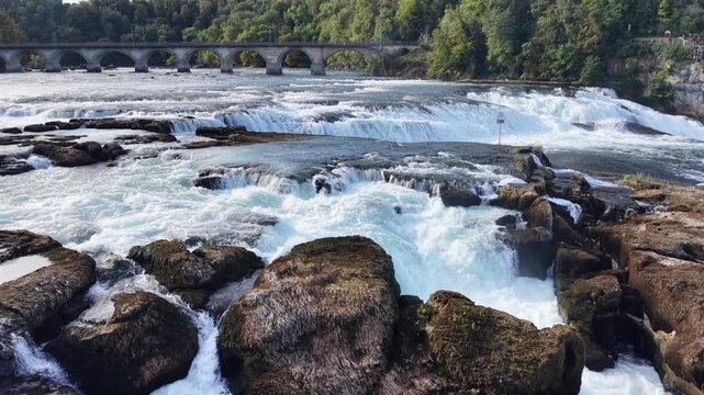 Close aerial view of Rhine Falls in Switzerland, showing powerful water channels rushing between massive exposed rocks with the historic railway bridge in the background.