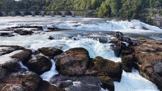 Close aerial view of Rhine Falls in Switzerland, showing powerful water channels rushing between massive exposed rocks with the historic railway bridge in the background.