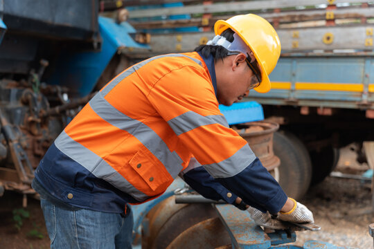 Side view of Asian male technician in yellow hard hat and reflective jacket checking and repairing heavy machinery component outdoors.
