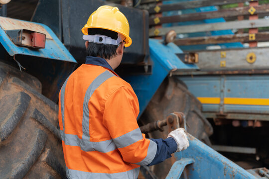 Back view of male worker wearing yellow hard hat and orange safety vest inspecting heavy machinery vehicle on industrial construction site.