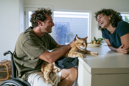 Cat wearing witch hat perched on a mans armrest in a kitchen, with a laughing housemate for pet retail and accessibility campaigns