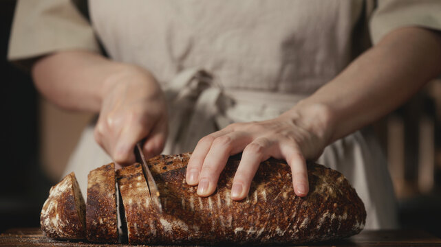 Close-up of baker in rustic linen apron slicing freshly baked artisan sourdough bread with knife on wooden cutting board. Homemade crusty loaf for breakfast, bakery promos, and healthy food concepts.
