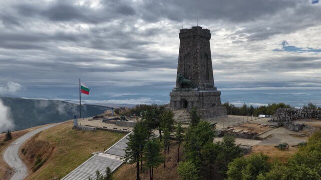 Aerial drone view of the Shipka Monument in Bulgaria, rising proudly above the Balkan Mountains with a Bulgarian flag, historic cannons and dramatic clouds surrounding the mountain peak.