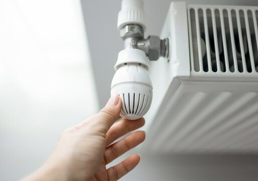 Hands adjusting a white radiator thermostat and feeling the heater. The set includes 50 Euro banknotes and a black wallet placed on the heating equipment.