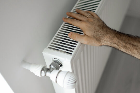 Hands adjusting a white radiator thermostat and feeling the heater. The set includes 50 Euro banknotes and a black wallet placed on the heating equipment.
