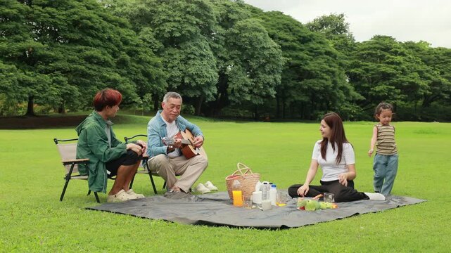 Asian kids painting in park. Asian family enjoy outdoor picnic with food and drinks in park sunny day, outdoor activitie with daughter father, mother grandfather. GrandFather playing acoustic guitar