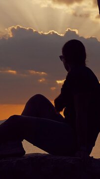 Vertical video. Silhouette of Young Woman Adjusting Position While Sitting on Large Rock Watching Sunset Over Calm Sea, Peaceful Evening, Coastal Lifestyle, Travel, Relaxation