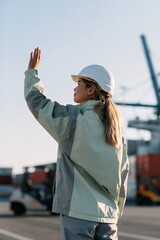 Fototapeta premium Female dock worker signaling at shipping port with safety helmet and jacket for logistics banner background and industrial transport design