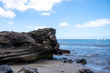 Coastal cliffs meet calm blue waters under clear sky. Sailboats drift far on horizon. Rocky shore shows weathered textures. Serene scene invites relaxation or travel inspiration © AlbertoRodriguez