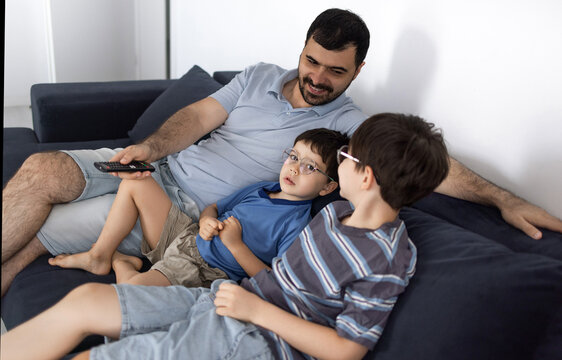 Father and his two sons sitting on a couch and watching television in a living room. The man holds a remote control while relaxing with the boys indoors
