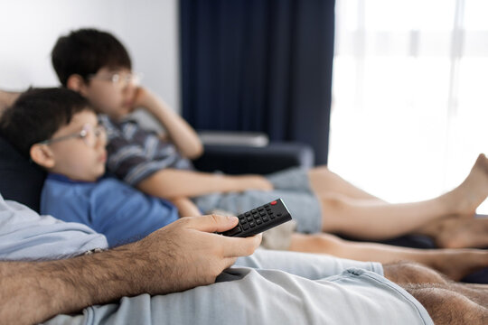 Father and his two sons sitting on a couch and watching television in a living room. The man holds a remote control while relaxing with the boys indoors
