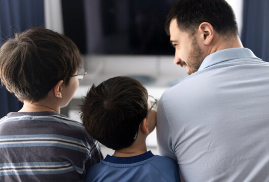 Father and his two sons sitting on a couch and watching television in a living room. The man holds a remote control while relaxing with the boys indoors