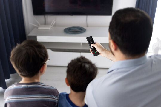 Father and his two sons sitting on a couch and watching television in a living room. The man holds a remote control while relaxing with the boys indoors