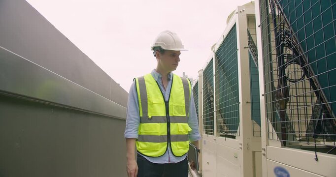 A focused HVAC technician wearing safety gear inspects a large commercial air conditioning unit on a building rooftop during a routine maintenance check.