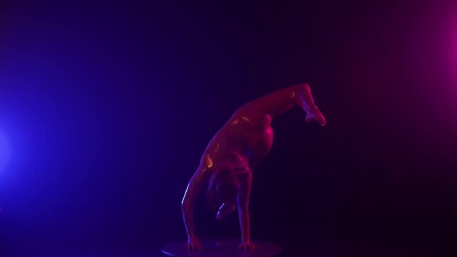 Female acrobat in a shiny bodysuit performs a dramatic handstand split on a pedestal, illuminated by vibrant blue and magenta studio lights on a dark stage.