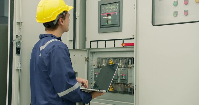 An electrical engineer performs a system diagnostic check with a laptop, conducting maintenance on an industrial power distribution control panel in a factory.