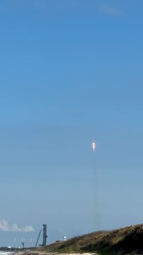 A rocket booster returns through a clear blue sky toward a coastal spaceport near the ocean. The landing burn, smoke trail, launch tower, beach, and dunes show a dramatic reusable rocket recovery
