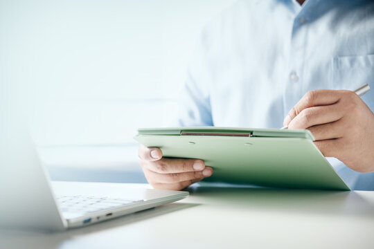 Businessman writing notes on a green clipboard while working on a laptop at office desk, concept of corporate administration, planning, task management, and professional documentation.